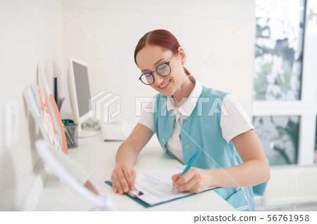 Red-haired dentist wearing glasses filling in some form Red-haired dentist wearing glasses filling in some form 56761958