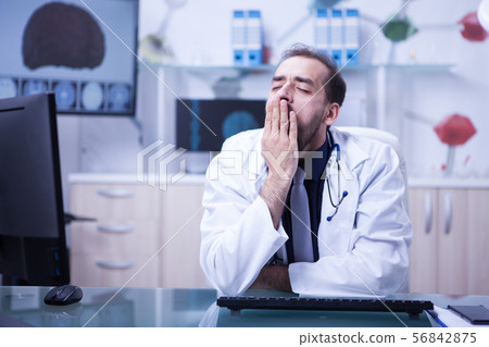 Young bearded doctor tired and yawning sitting at the office in his hospital cabinet Young bearded doctor tired and yawning sitting at the office in his hospital cabinet 56842875
