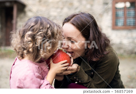 A small girl with mother standing in front of old stone house, holding pumpkin. 56857169