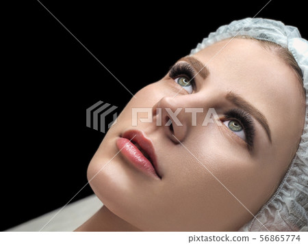 Eyelash extension result. Female face with long black lashes and disposable cap on couch in beauty 56865774