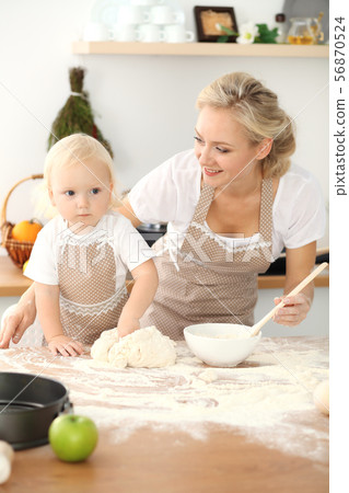 Little girl and her blonde mom in beige aprons playing and laughing while kneading the dough in Little girl and her blonde mom in beige aprons playing and laughing while kneading the dough in 56870524