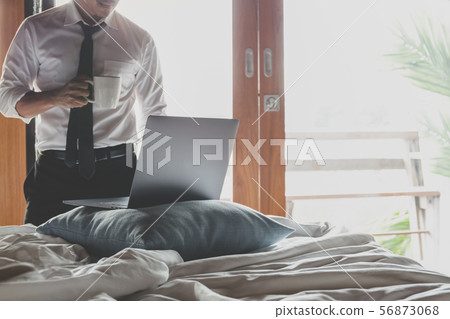 Businessman holding cup of coffee and working on laptop in bed room. Businessman holding cup of coffee and working on laptop in bed room. 56873068