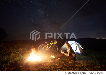 Woman resting at night camping near campfire, tourist tent, bicycle under evening sky full of stars 56874598
