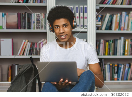 African american student with headset leaning on bookshelf, using laptop 56882022