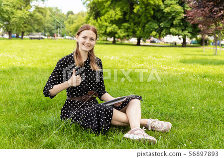 Young blonde girl in a summer dress is using a tablet and showing a thumbs up sign while sitting on Young blonde girl in a summer dress is using a tablet and showing a thumbs up sign while sitting on 56897893