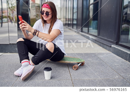 Photo of happy brunette in sunglasses with phone in her hands sitting on skateboard on background of Photo of happy brunette in sunglasses with phone in her hands sitting on skateboard on background of 56976165