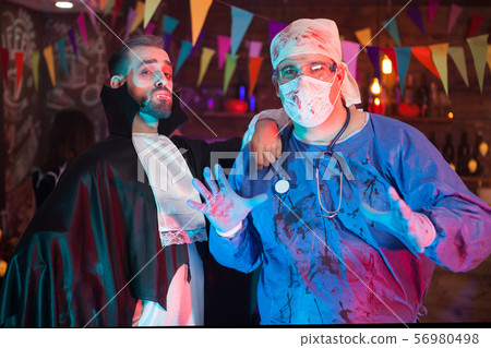 Scary man in a doctor costume covered in blood with his handsome dracula friend celebrating Scary man in a doctor costume covered in blood with his handsome dracula friend celebrating 56980498