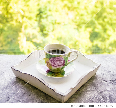 Hot morning coffee in small floral design cup on shabby chic vintage white tray on window sill in Hot morning coffee in small floral design cup on shabby chic vintage white tray on window sill in 56985289