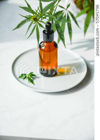 CBD Oil bottle and dropper on a small plate and on a marble surface. Cannabis leaves nearby. 57045796