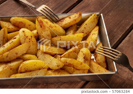Potato wedges, oven baked, in a baking tray on a dark rustic wooden background, with two forks 57063284