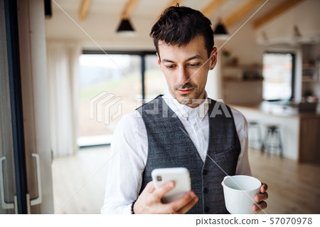 Young man with coffee and smartphone standing indoors at home. 57070978