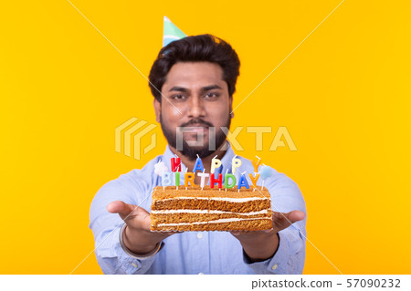 Positive funny young indian guy with a cap and a homemade cake in his hands posing on a yellow Positive funny young indian guy with a cap and a homemade cake in his hands posing on a yellow 57090232