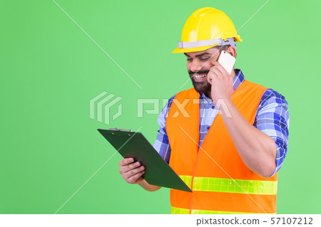 Happy young overweight bearded Indian man construction worker reading on clipboard and talking on Happy young overweight bearded Indian man construction worker reading on clipboard and talking on 57107212