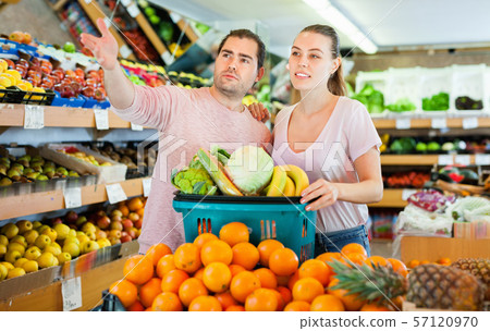 Positive family couple standing with full cart after shopping and pointing to shelves in fruit store Positive family couple standing with full cart after shopping and pointing to shelves in fruit store 57120970