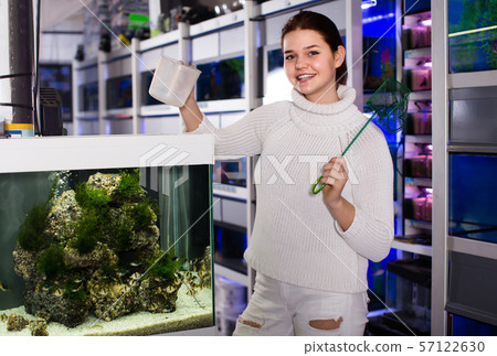 Girl holding aquarian net and water container next to aquarium with colorful fish in aquarium shop 57122630