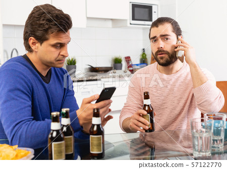 Two concerned men talking and laughing while enjoying beer at home, using phone Two concerned men talking and laughing while enjoying beer at home, using phone 57122770