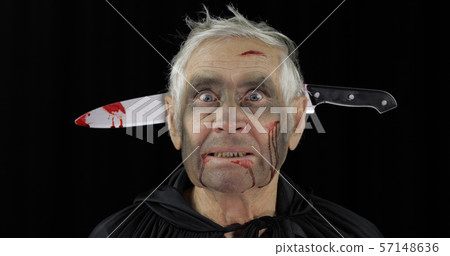 Elderly man with knife in head. Halloween makeup and costume. Blood on his face Elderly man with knife in head. Halloween makeup and costume. Blood on his face 57148636