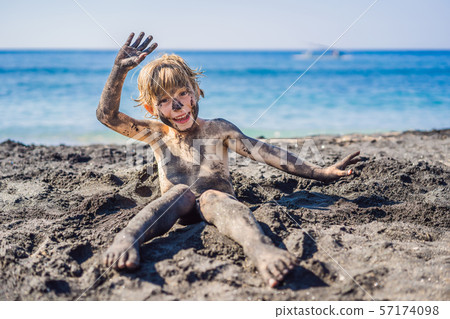 Black Friday concept. Smiling boy with dirty Black face sitting and playing on black sand sea beach Black Friday concept. Smiling boy with dirty Black face sitting and playing on black sand sea beach 57174098