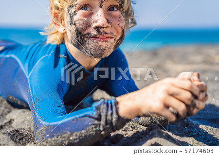 Black Friday concept. Smiling boy with dirty Black face sitting and playing on black sand sea beach Black Friday concept. Smiling boy with dirty Black face sitting and playing on black sand sea beach 57174603