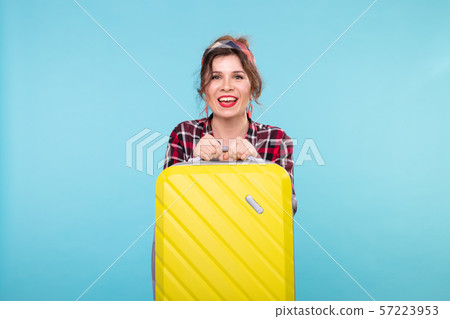 Positive young smiling woman in a plaid shirt holding a yellow suitcase posing on a blue background Positive young smiling woman in a plaid shirt holding a yellow suitcase posing on a blue background 57223953