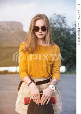 Beautiful and fashionable young woman in sunglasses and with a tattoo poses with a skateboard or 57243003
