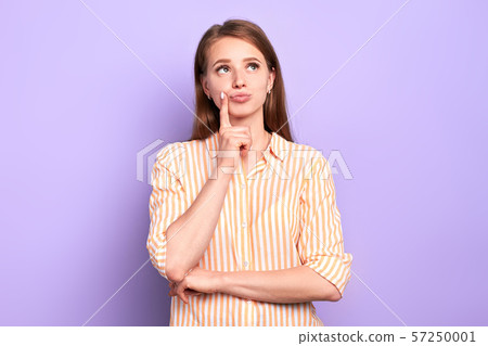 Dreamy pretty female keeps index finger near lips, looking somewhere above 57250001