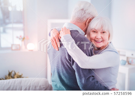 Beaming retired woman feeling happy hugging her grey-haired husband Beaming retired woman feeling happy hugging her grey-haired husband 57258596