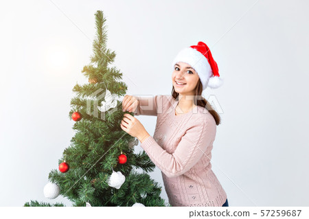 Holidays and celebration concept - Happy brunette woman in santa's hat decorates a christmas tree at 57259687