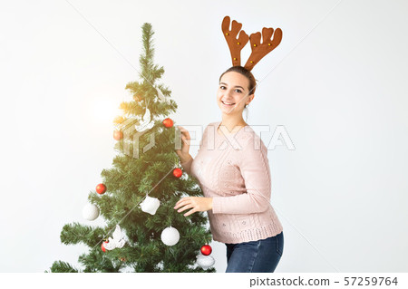 Holidays and celebration concept - Happy brunette woman in funny deer hat decorates a christmas tree Holidays and celebration concept - Happy brunette woman in funny deer hat decorates a christmas tree 57259764