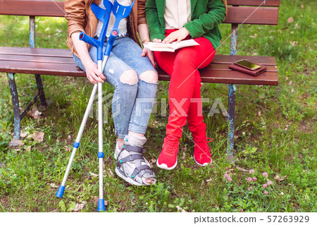 Two friends reading a book in the park, one has a broken foot 57263929