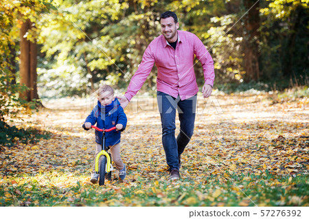 Father and small son with a balance bike on a walk in autumn forest. Father and small son with a balance bike on a walk in autumn forest. 57276392
