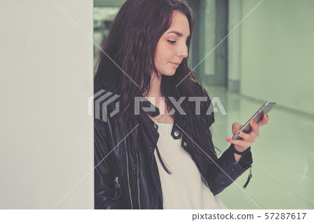 A beautiful girl uses a smartphone in an office building leaning against a wall A beautiful girl uses a smartphone in an office building leaning against a wall 57287617