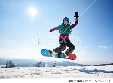 Female snowboarder jumping in air on background of blue sky and snowy mountains on sunny day. 57322491