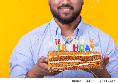 Close-up of funny young indian guy with a cap and a homemade cake in his hands posing on a yellow 57349587