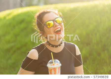 Portrait of a positive young pretty girl in punk clothes and glasses with a milkshake in her hands Portrait of a positive young pretty girl in punk clothes and glasses with a milkshake in her hands 57349639