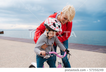 Mother helping small daughter to ride bicycle outdoors on beach. 57350249