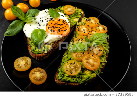 Open sandwiches with avocado guacamole, yellow cherry tomatoes, fried egg and basil on a black plate Open sandwiches with avocado guacamole, yellow cherry tomatoes, fried egg and basil on a black plate 57359593
