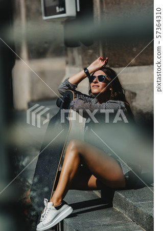 Portrait of Beautiful young woman sitting on the ground and holding a longboard in the city street 57364310