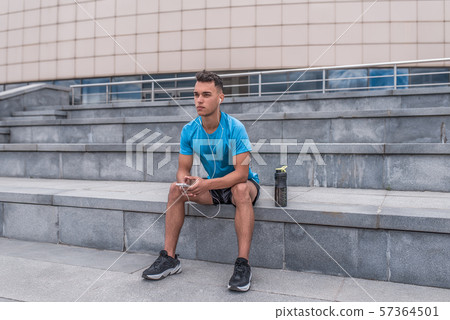 Athletic male athlete, summer city sitting hand, phone listens music headphones motivation fitness Athletic male athlete, summer city sitting hand, phone listens music headphones motivation fitness 57364501