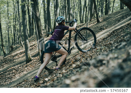 Image of female athlete in helmet raising bicycle to hill in forest Image of female athlete in helmet raising bicycle to hill in forest 57450167