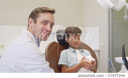Mature dentist smiling to the camera while working with adorable little boy Mature dentist smiling to the camera while working with adorable little boy 57475344