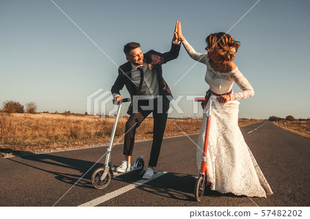 Smiling wedding couple riding a on scooters along the road outside the city at sunset. 57482202