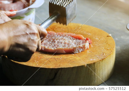 The man's hand picks up an iron hammer and smashed the pork on a wooden chopping board to make steak. The man's hand picks up an iron hammer and smashed the pork on a wooden chopping board to make steak. 57525512