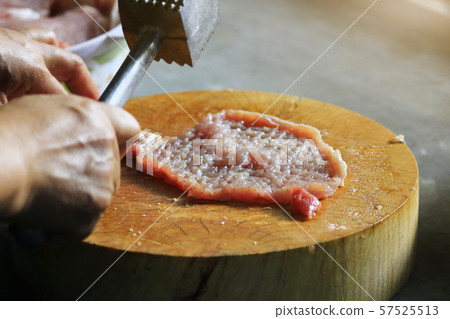 The man's hand picks up an iron hammer and smashed the pork on a wooden chopping board to make steak. The man's hand picks up an iron hammer and smashed the pork on a wooden chopping board to make steak. 57525513