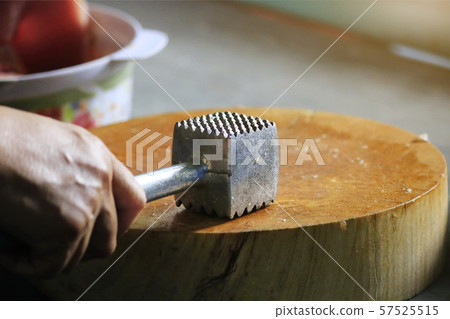 The man's hand picks up an iron hammer and smashed the pork on a wooden chopping board to make steak. The man's hand picks up an iron hammer and smashed the pork on a wooden chopping board to make steak. 57525515