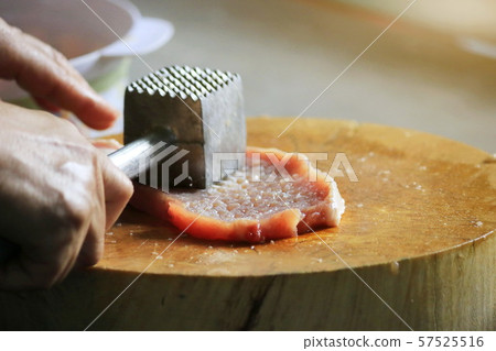 The man's hand picks up an iron hammer and smashed the pork on a wooden chopping board to make steak. The man's hand picks up an iron hammer and smashed the pork on a wooden chopping board to make steak. 57525516