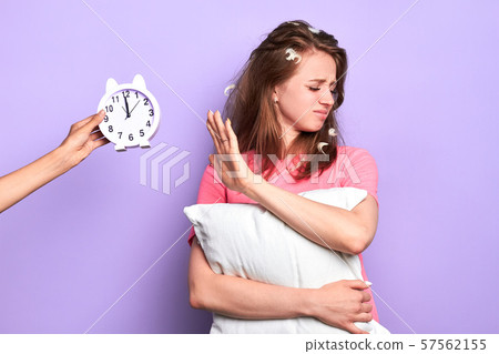 Dissatisfied young lady holds pillow, waves aside alarm clock 57562155