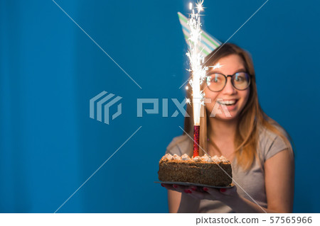 Cheerful young blurred girl student in glasses holding a congratulatory cake with a candle standing Cheerful young blurred girl student in glasses holding a congratulatory cake with a candle standing 57565966