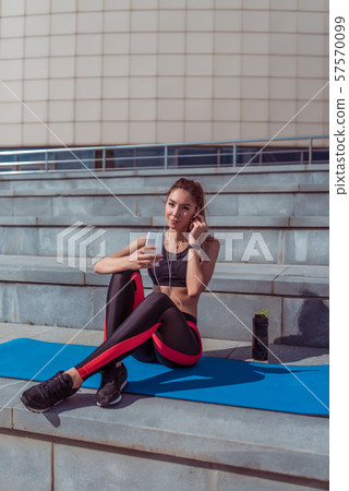 A beautiful woman athlete sits on a yoga mat, holds a phone, listens to music on headphones, a 57570099