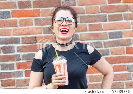 Young positive girl holds in Ruach a milkshake with a straw against a brick wall background. Summer 57589876